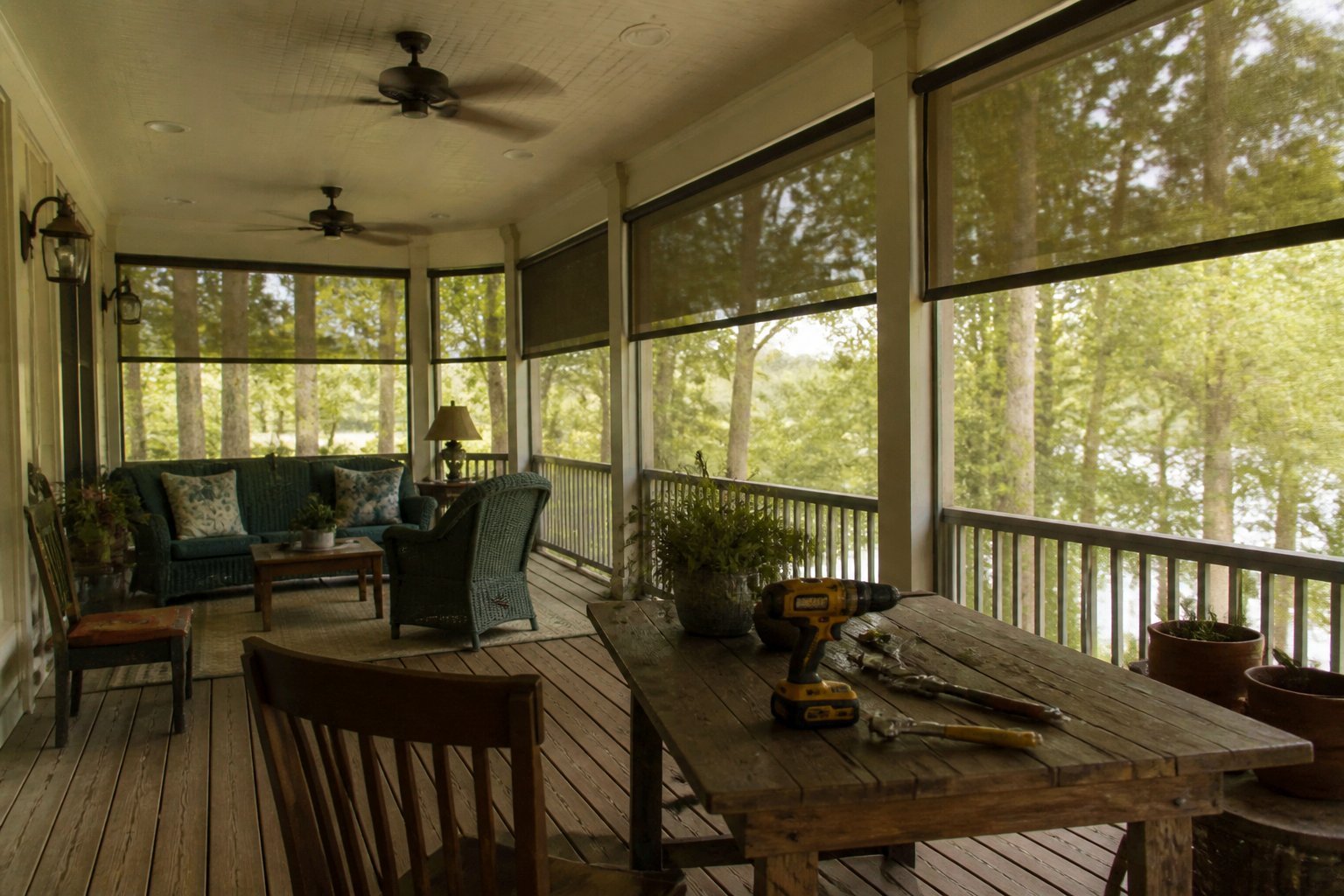 A screened wrap-around porch with overhead fans and partially lowered retractable shades, showing a wooden table with gently used tools and objects in soft daylight.