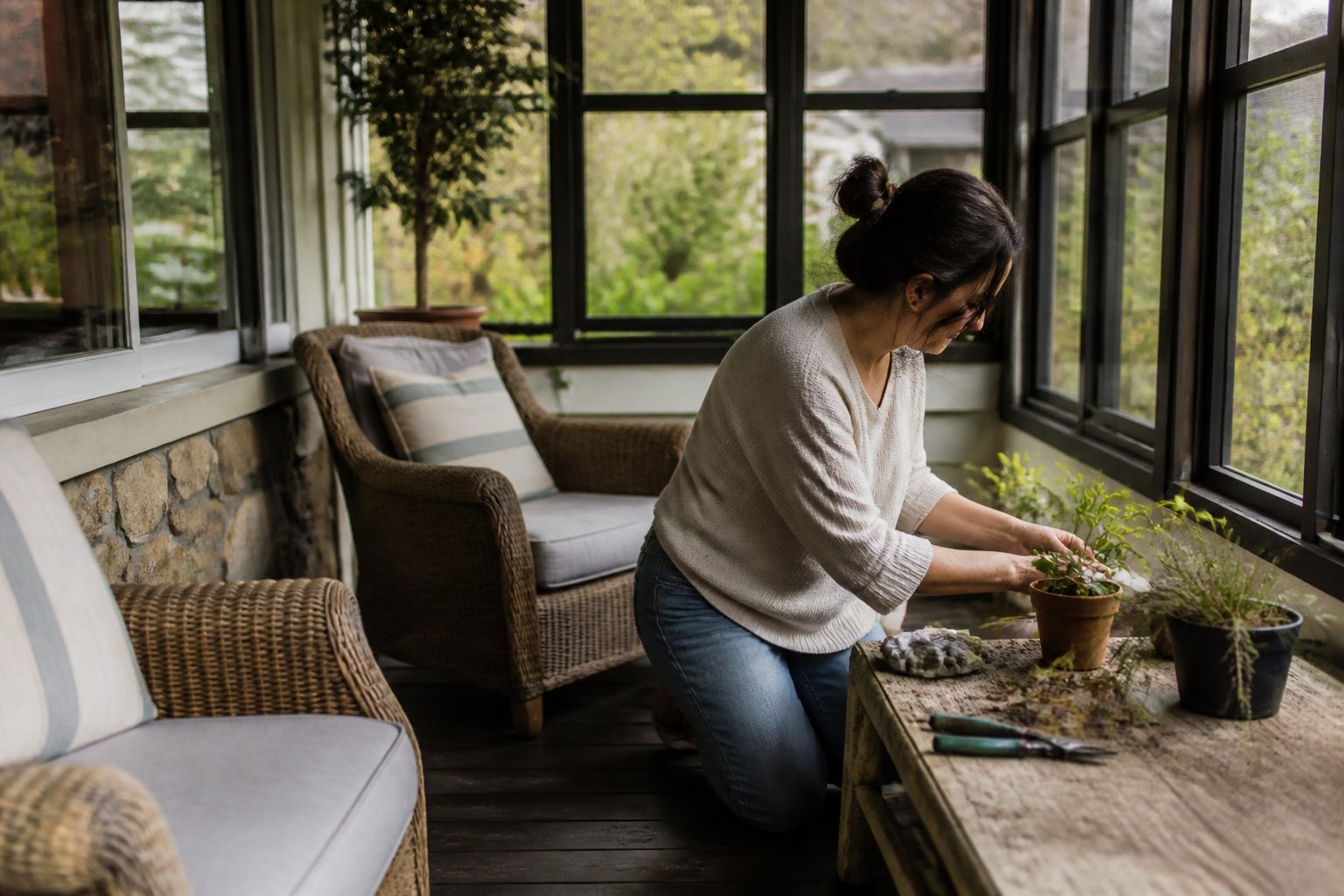 A cozy enclosed porch with seating, gardening tools, and hands arranging items, bathed in soft daylight with natural shadows.